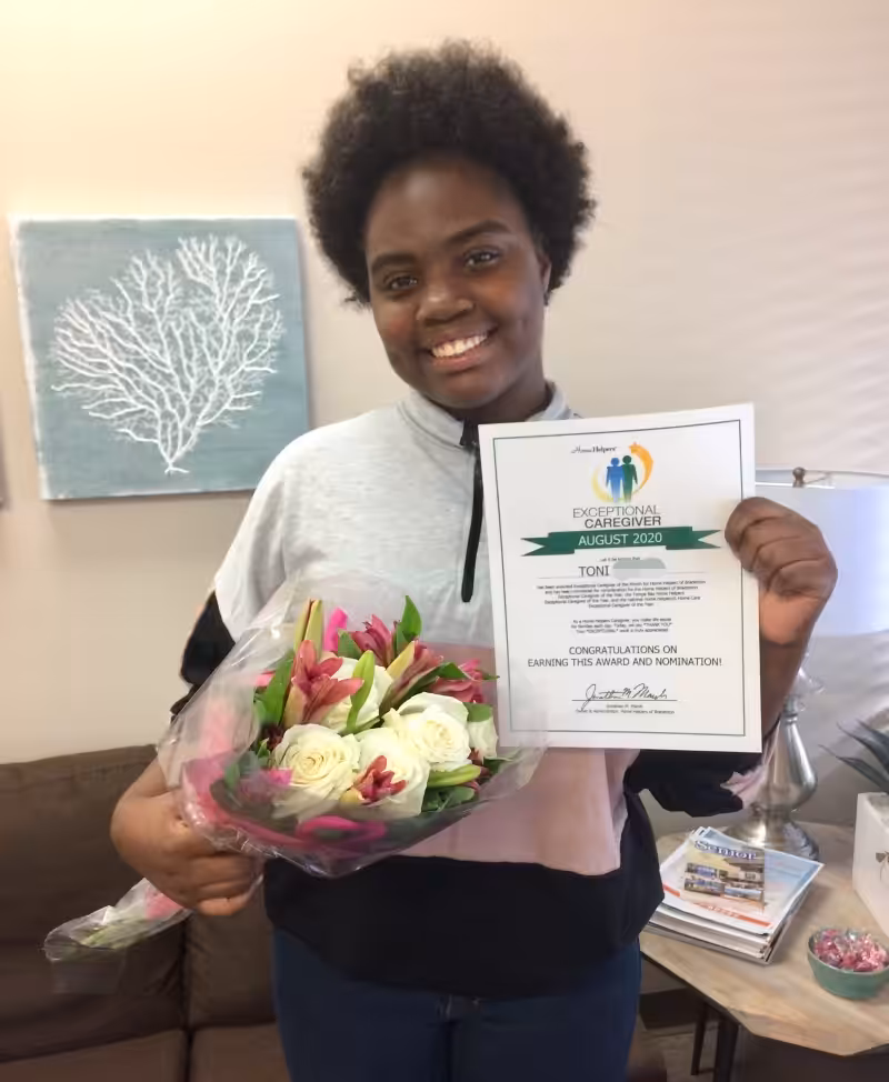 A young woman smiles while holding a bouquet of flowers in one hand and a certificate in the other. She stands indoors by a couch and a wall with art, celebrating her achievement.