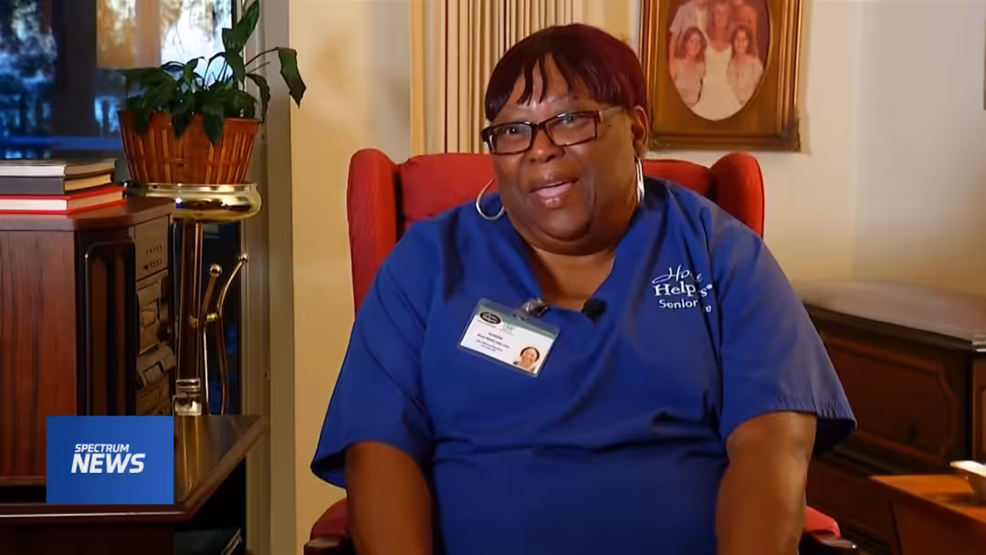 A woman in blue scrubs with a name badge sits in a red chair, smiling. Behind her are family photos, a plant, and a bookshelf. A Spectrum News logo appears in the lower left corner.