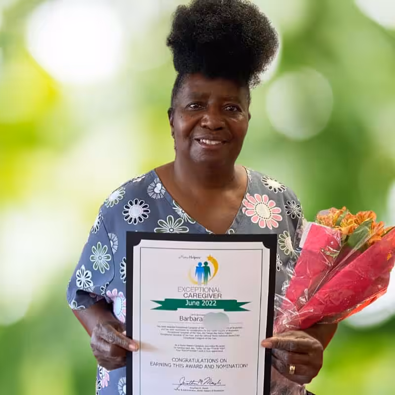 A smiling woman in floral scrubs holds a bouquet of flowers and a framed Exceptional Caregiver June 2022 award, standing in front of a blurred green background.