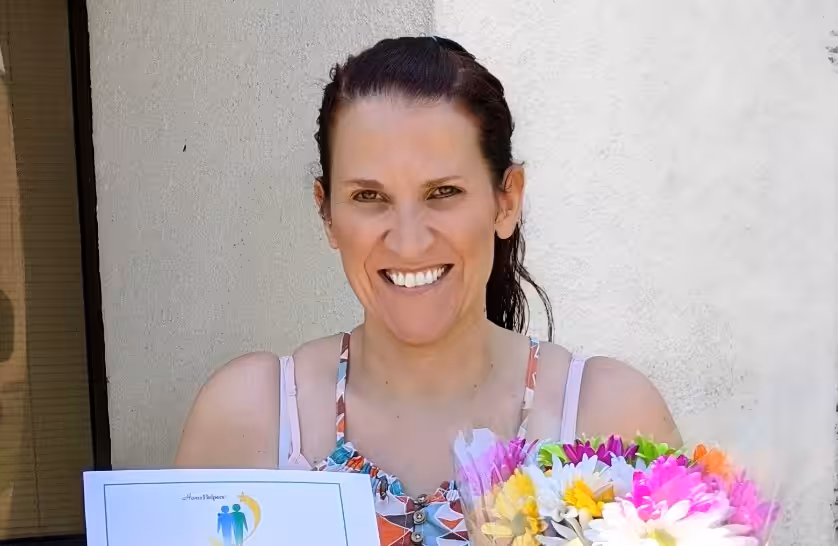 A smiling woman with brown hair in a ponytail holds a bouquet of colorful flowers and a certificate while standing in front of a light-colored wall.