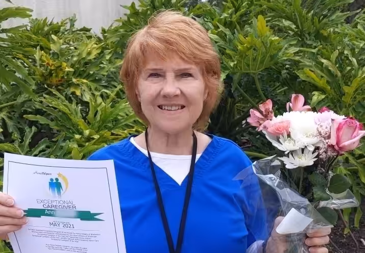 A smiling woman with short red hair holds a bouquet of flowers and a certificate titled Exceptional Caregiver Award in front of green plants. She is wearing blue scrubs and a lanyard.