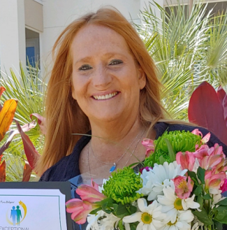 A smiling woman with long red hair holds a bouquet of colorful flowers and a certificate, standing outdoors with palm leaves and red plants in the background.