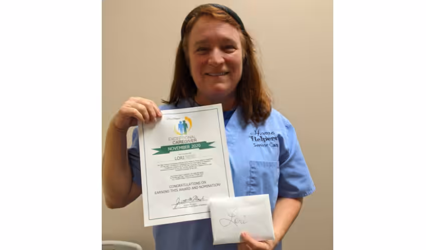 A smiling woman in a blue Home Helpers Senior Care shirt holds a certificate for Exceptional Caregiver November 2020 and an envelope labeled Lori against a plain indoor background.
