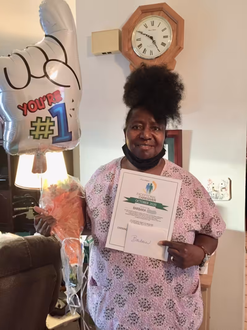 A woman in a pink scrub top holds a Youre #1 balloon, a bouquet of flowers, and a certificate, smiling. A wall clock and lamp are visible in the background.