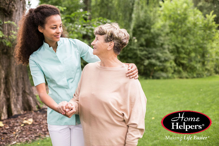 A young caregiver smiles and gently holds hands with an elderly woman as they walk together outside in a green park. The Home Helpers logo and the slogan Making Life Easier are visible in the bottom right corner.