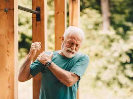 An older man in a green shirt stands outdoors, holding his shoulder and wincing in pain, possibly from an injury, next to a wooden pull-up bar structure surrounded by greenery.