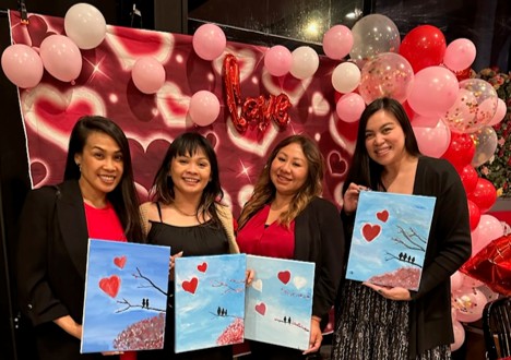 Four women stand in front of a heart and balloon-filled backdrop with a “love” sign, each holding a painting of birds on a branch with red hearts in the sky. All are smiling and dressed in black and red.