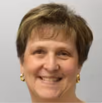 A woman with short light brown hair smiles at the camera. She is wearing gold earrings and a light-colored top, with a plain, light gray background behind her.