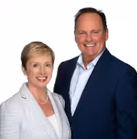 A smiling woman and man pose together in professional attire against a white background. The woman wears a light-colored blazer and the man wears a dark blue suit jacket and white shirt.
