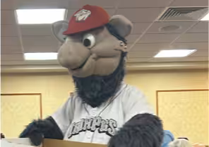 A person in a furry animal mascot costume wearing a red cap and a white jersey with Stripers written on it stands indoors under a ceiling with fluorescent lights.