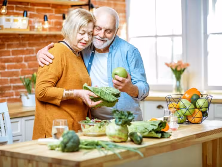 An elderly couple stands together in a bright kitchen, smiling as they prepare vegetables. The woman holds leafy greens, and the man holds an apple. A basket of fruit and vegetables sits on the counter nearby.