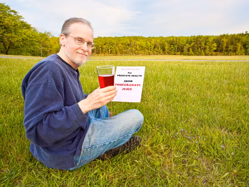 A man sitting on grass outdoors, holding a glass of red juice. He smiles at the camera. Beside him is a sign that reads, For Prostate Health, Drink Pomegranate Juice. Trees and open fields are in the background.