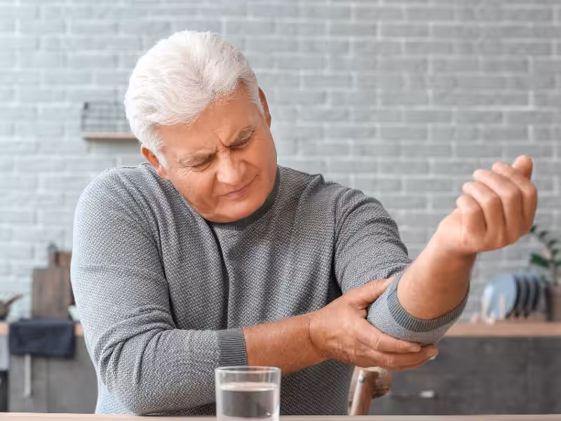 An older man with gray hair sits at a table, grimacing in pain as he holds his left elbow with his right hand. A glass of water is on the table in front of him. The background shows a kitchen setting.