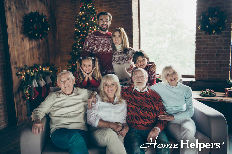 A multigenerational family sits together on a couch and stands behind it, smiling in a festive living room decorated with Christmas trees and stockings. The Home Helpers logo is in the bottom right corner.