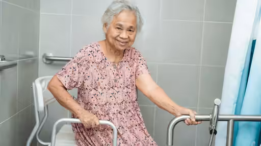 An elderly woman sits in a shower chair, holding onto grab bars in a tiled bathroom, smiling at the camera. She is wearing a pink patterned dress.