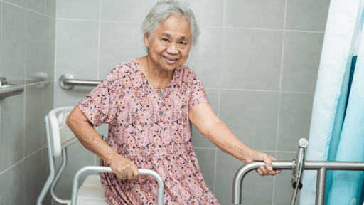 An elderly woman sits in a shower chair, holding onto grab bars in a tiled bathroom, smiling at the camera. She is wearing a pink patterned dress.