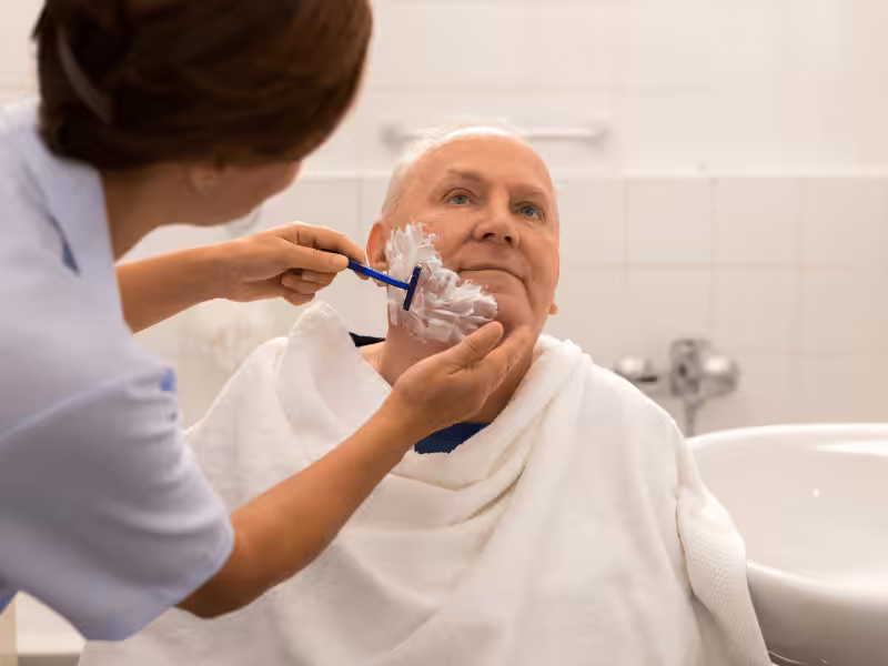 A caregiver is gently shaving an elderly man’s face with a razor and shaving cream while he sits in a chair, covered with a white towel, in a bright bathroom setting.