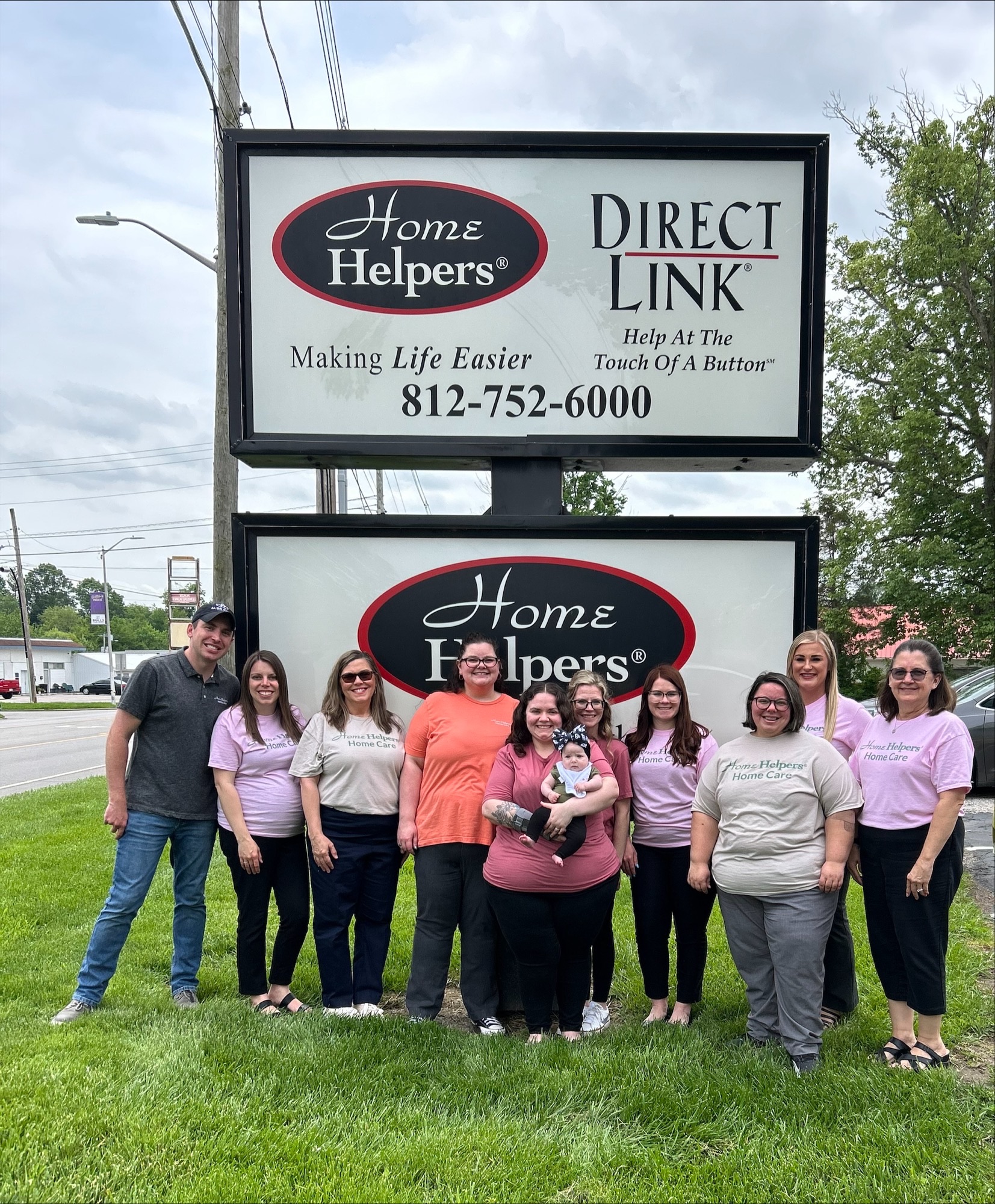 A group of eleven adults, some in matching shirts, stand and smile in front of a large Home Helpers and Direct Link sign on a grassy area by the road. One person in the middle holds a small dog.