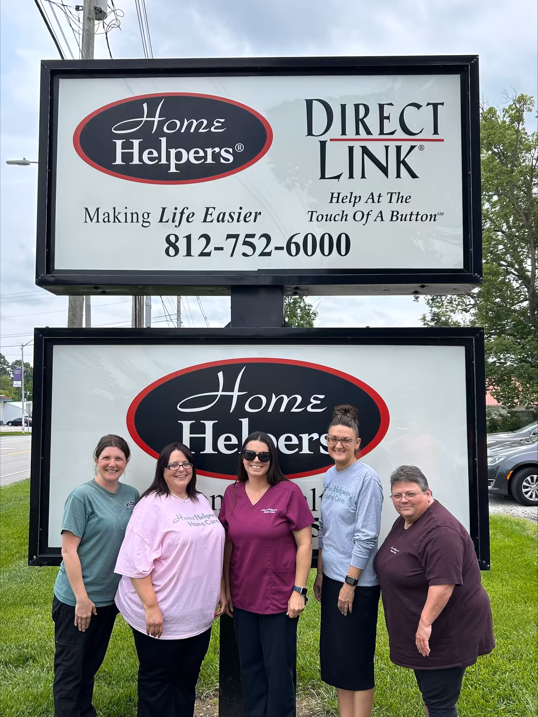 Five women stand smiling in front of two business signs for Home Helpers and Direct Link displaying a phone number, outside on a cloudy day with grass and trees in the background.