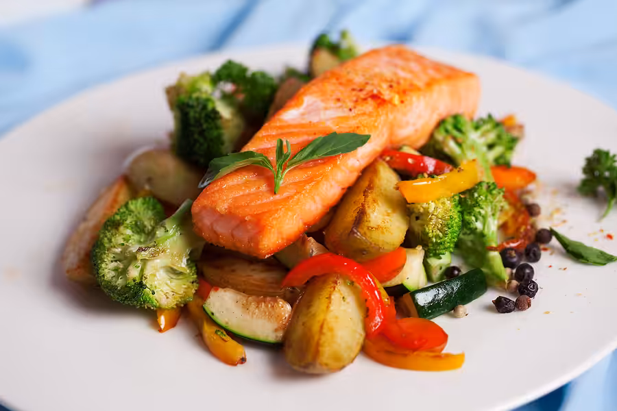 A plate with a cooked salmon fillet on top of roasted potatoes, broccoli, red and yellow bell peppers, zucchini, and garnished with fresh herbs.