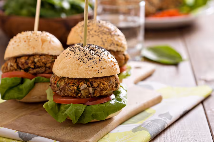 Two veggie burgers with lettuce and tomato on poppy seed buns are served on a wooden board. A glass of water and a leafy green garnish are visible in the background.