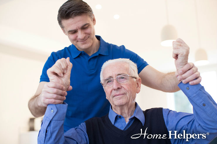 A younger man assists an elderly man with arm exercises at home. Both appear focused; the younger man is supportive, standing behind the older man. The image features the “Home Helpers” logo in the bottom right corner.