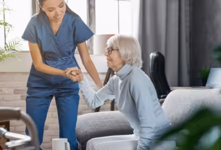 A caregiver in blue scrubs helps an elderly woman with gray hair and glasses stand up from a couch in a bright, comfortable living room.
