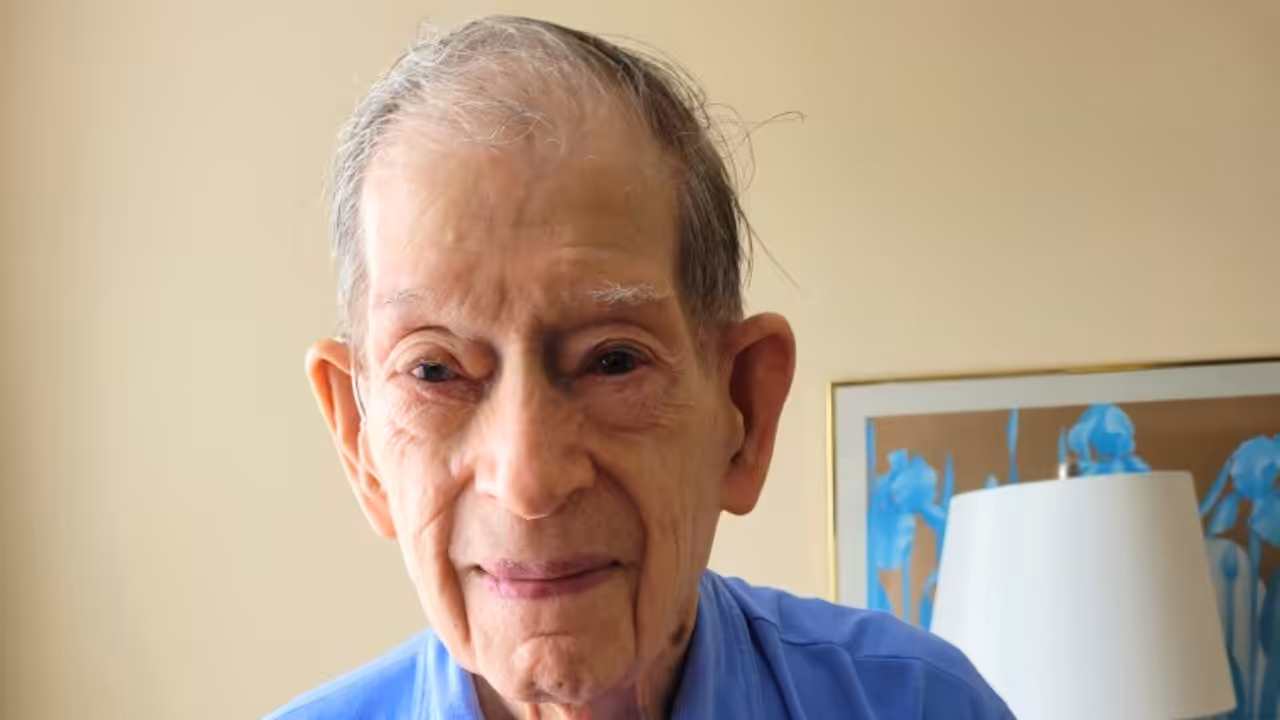 An elderly man with thinning gray hair and a gentle smile wears a light blue collared shirt. He stands indoors in front of a beige wall, with a framed picture and lamp in the background.