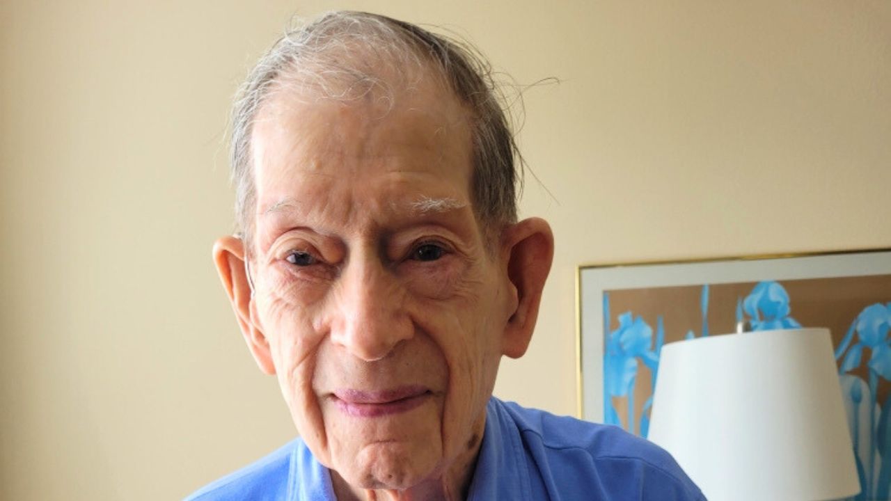 An elderly man with thinning gray hair and a gentle smile wears a light blue collared shirt. He stands indoors in front of a beige wall, with a framed picture and lamp in the background.