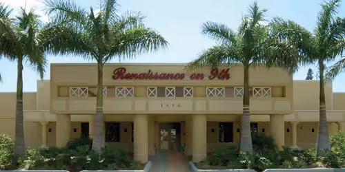 Beige building with Renaissance on 9th written in red script above the entrance, surrounded by palm trees and greenery, under a clear blue sky.