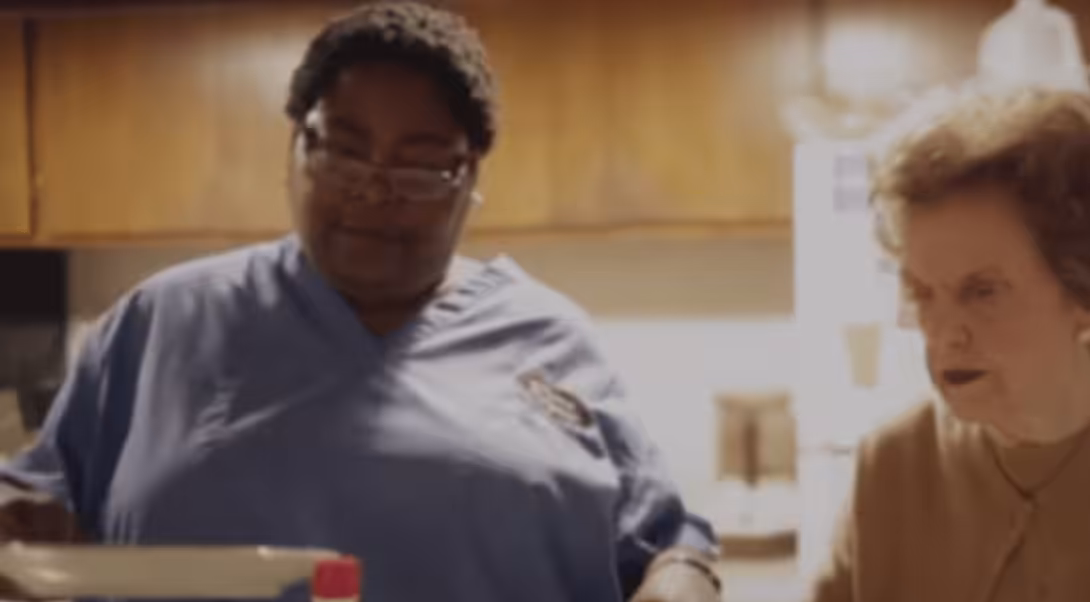 A woman in blue scrubs stands next to an older woman in a kitchen, both focused on a task out of view. The background shows cabinets and kitchen appliances.