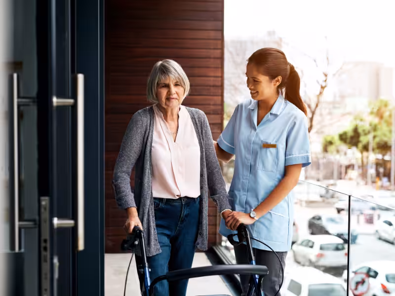 A caregiver in a light blue uniform assists an older woman using a walker on a balcony. Both are smiling, and parked cars and trees are visible in the background.