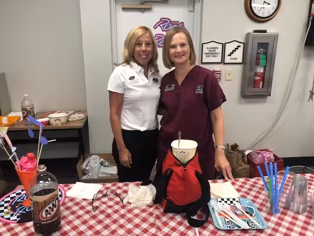 Two women stand behind a checkered table with snacks, sodas, plates, and decorations. One wears a white shirt and the other wears maroon scrubs. They are smiling in what looks like an indoor event or office setting.