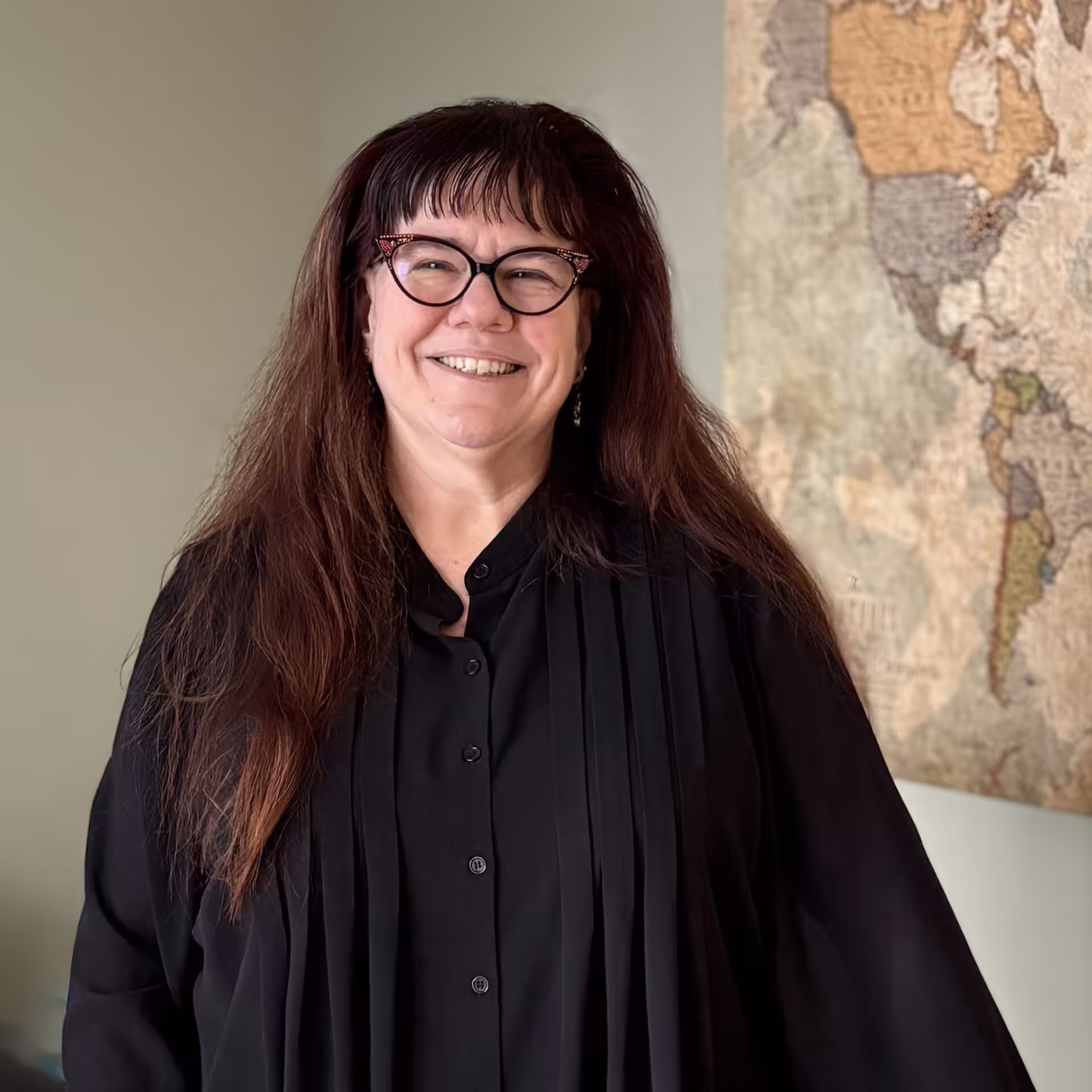 A smiling woman with long dark hair and glasses wears a black shirt and stands indoors near a wall with a vintage world map.
