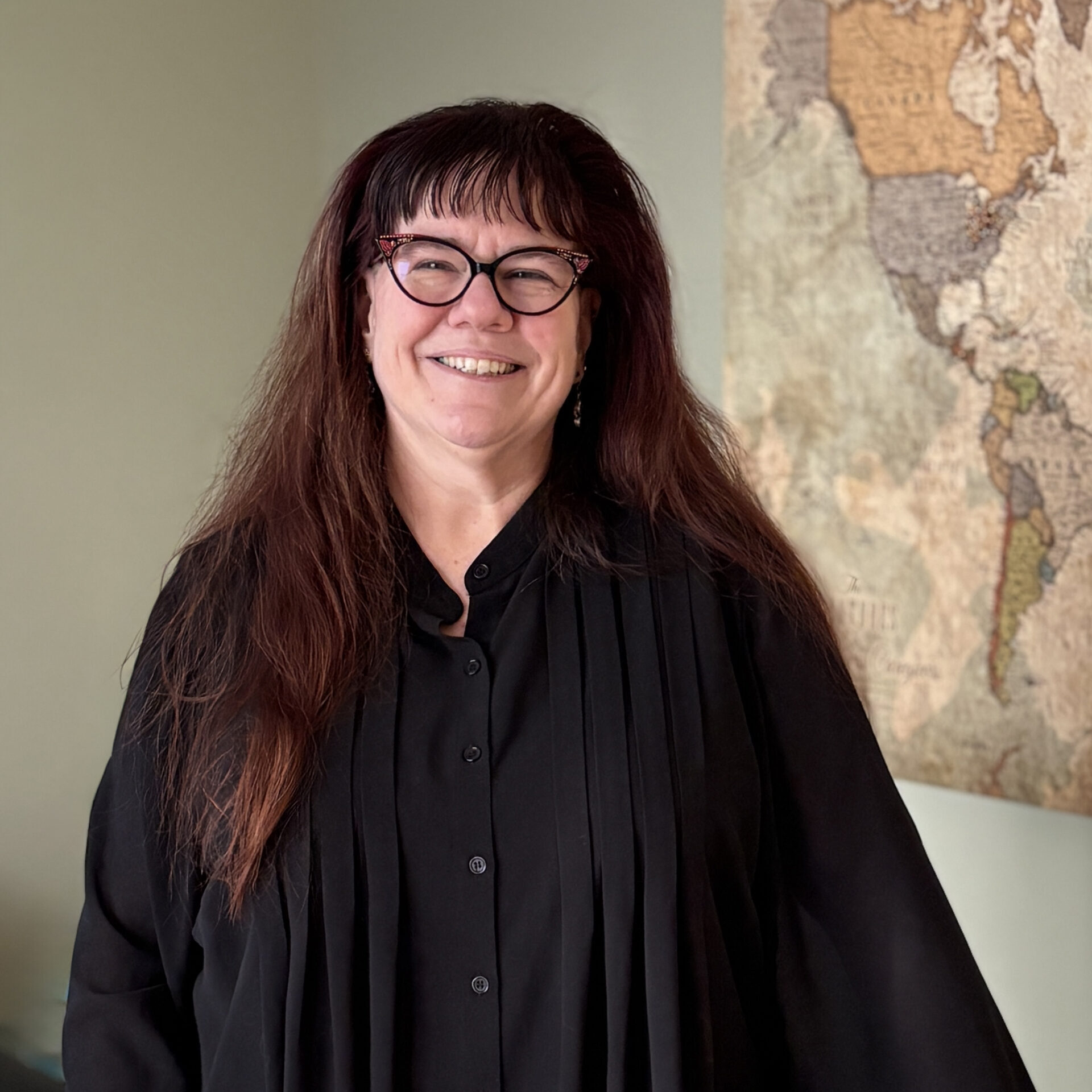 A smiling woman with long dark hair and glasses wears a black shirt and stands indoors near a wall with a vintage world map.