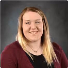 A woman with straight, blonde hair, wearing a burgundy sweater over a black top, smiles at the camera against a dark, neutral background.