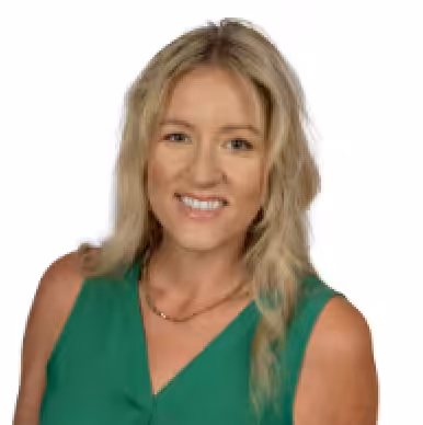 A smiling woman with wavy blonde hair wears a sleeveless green top and a gold necklace, posing against a white background.