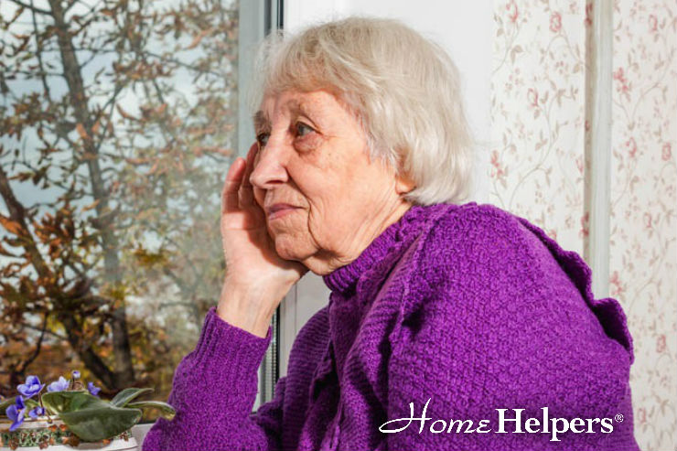 An elderly woman with short gray hair, wearing a purple sweater, sits by a window with a thoughtful expression. The Home Helpers logo appears in the lower right corner.