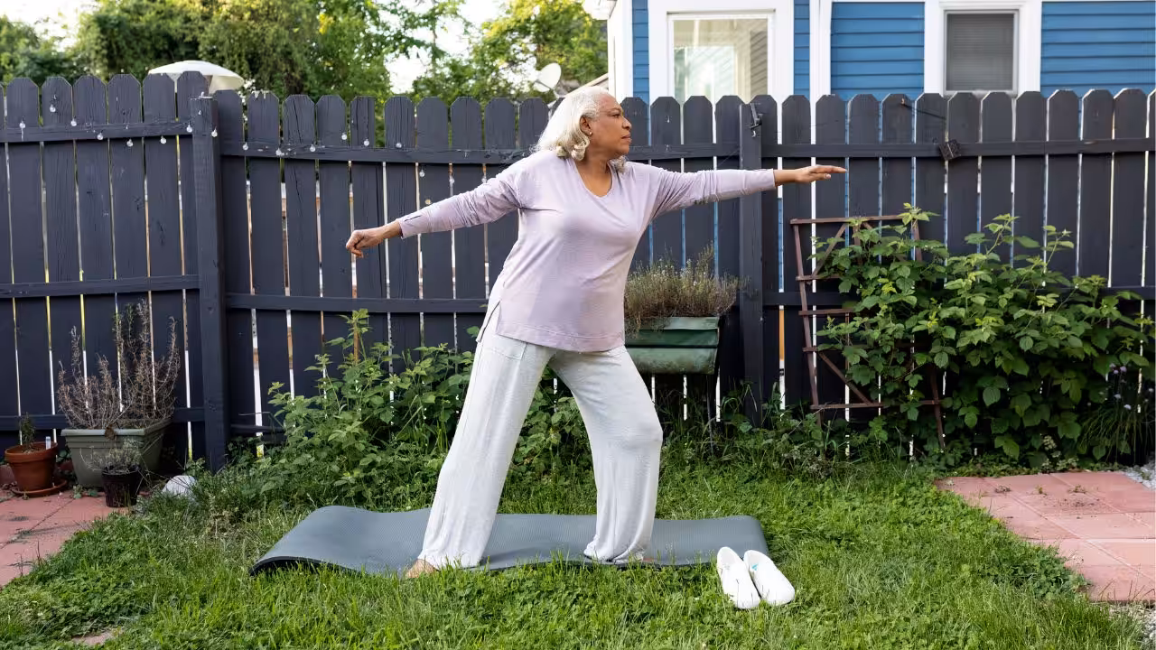 An older woman practices yoga on a mat in her backyard, standing in a warrior pose with arms outstretched. She wears comfortable clothes, and white slippers are placed beside the mat on the grass.