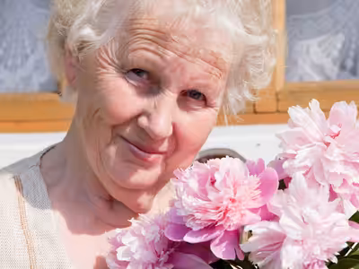 An elderly woman with short white hair smiles while holding a bouquet of pink flowers. She is standing outdoors with a window and lace curtains in the background.