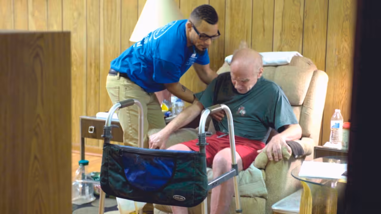 A younger man in a blue shirt assists an older man, seated in a recliner, as he stands using a walker in a wood-paneled room with a lamp, water bottle, and table beside them.