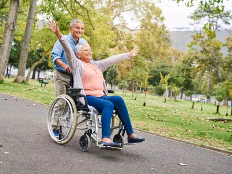 An older woman in a wheelchair, smiling with arms outstretched, is pushed by an older man along a paved path in a park surrounded by green trees. Both appear happy and are enjoying the outdoors.
