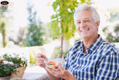 An older man with gray hair, wearing a blue plaid shirt, smiles while eating a bowl of fruit salad outdoors on a sunny day. Greenery and potted plants are visible in the background.