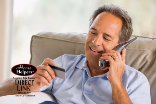 A smiling middle-aged man sits on a couch, holding a phone to his ear with one hand and a credit card in the other. The Home Helpers Direct Link logo appears in the lower left corner.