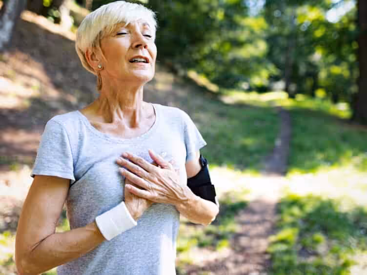 An older woman with short blonde hair stands outdoors on a wooded path, eyes closed and hand on her chest, appearing to catch her breath. She wears a light gray shirt and a fitness armband.
