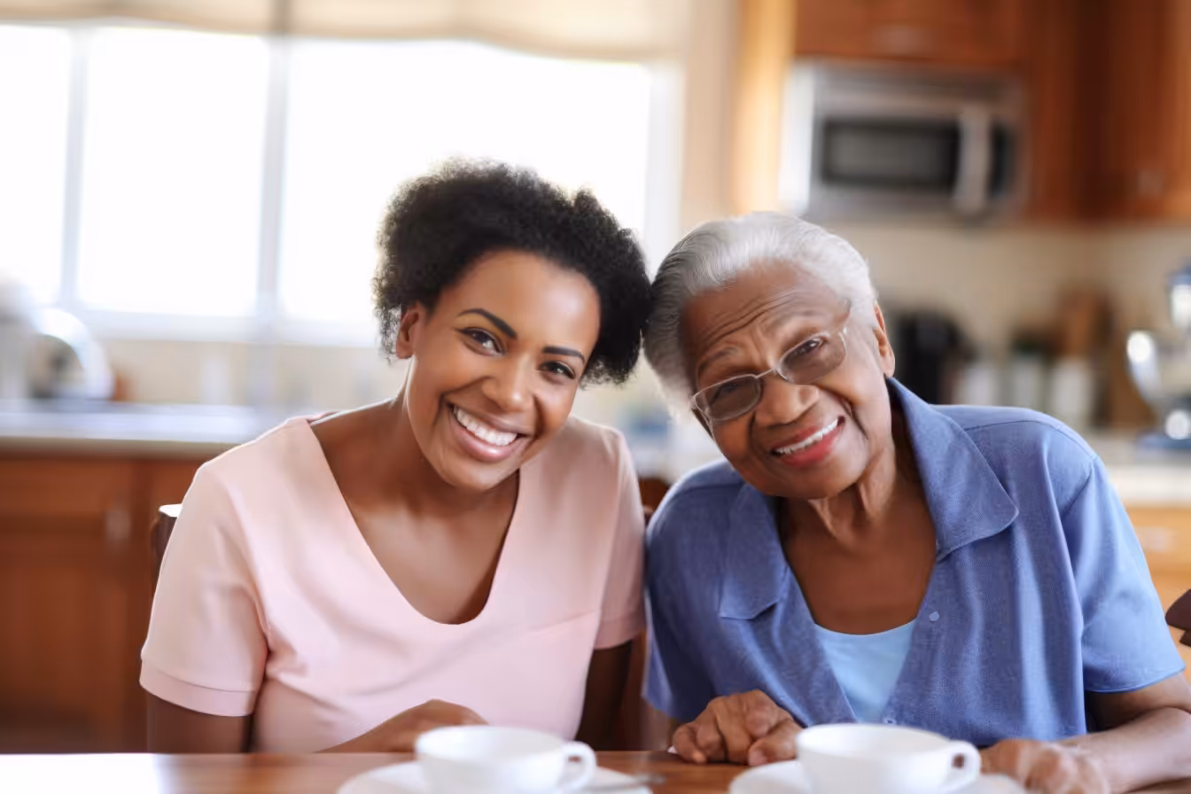 Two women, one younger and one older, sit close together at a kitchen table, both smiling warmly at the camera. There are teacups in front of them, and the background shows a brightly lit kitchen.