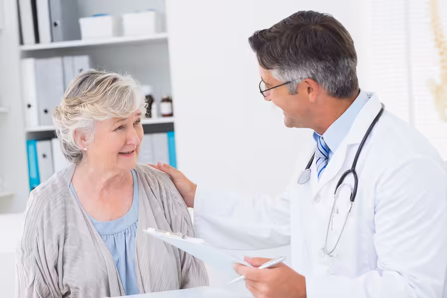 A doctor in a white coat smiles and talks with an older woman, gently touching her shoulder. The woman looks reassured and is sitting in a medical office with shelves and folders in the background.