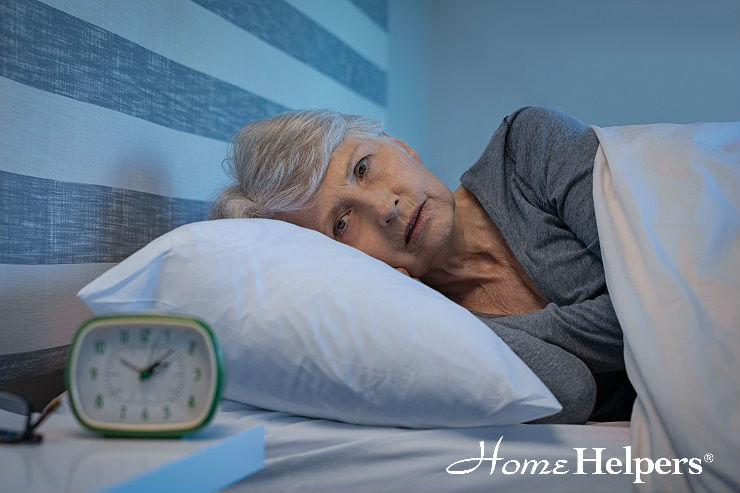 An elderly woman lies in bed awake at night, resting her head on a pillow and looking thoughtful. An alarm clock showing 4:10 sits on the bedside table. The Home Helpers logo appears in the lower right corner.
