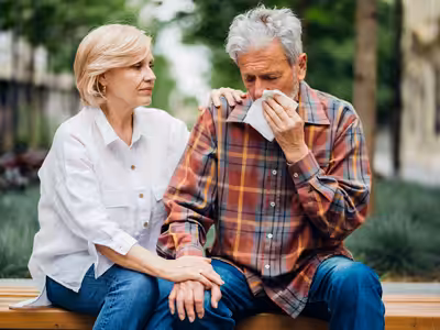 An older woman comforts an older man who is sitting on a bench, holding a tissue to his face as if he is crying. She rests one hand on his shoulder and the other on his hand, showing support outdoors.