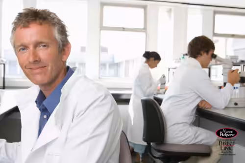 A man in a white lab coat smiles at the camera in a bright laboratory, while two other scientists work in the background, one using a microscope and the other handling lab equipment.